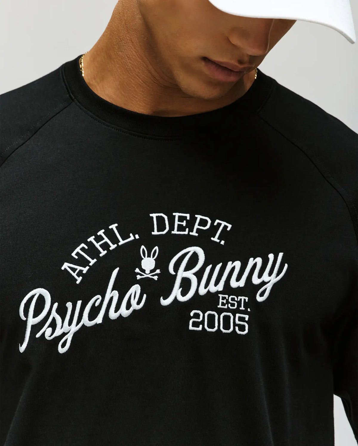 A man wears a Psycho Bunny MENS MATTEO RAGLAN GRAPHIC TEE (MT0200008) with white "ATHL. DEPT. Psycho Bunny EST. 2005" text and bunny logo, paired with a white cap; only his lower face is shown.
