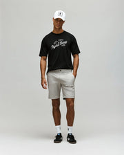 A man stands against a plain background wearing the Psycho Bunny MENS MATTEO RAGLAN GRAPHIC TEE (MT0200008), a white cap, light beige shorts, white socks, and black sandals. His arms are relaxed by his sides as he looks slightly downward.