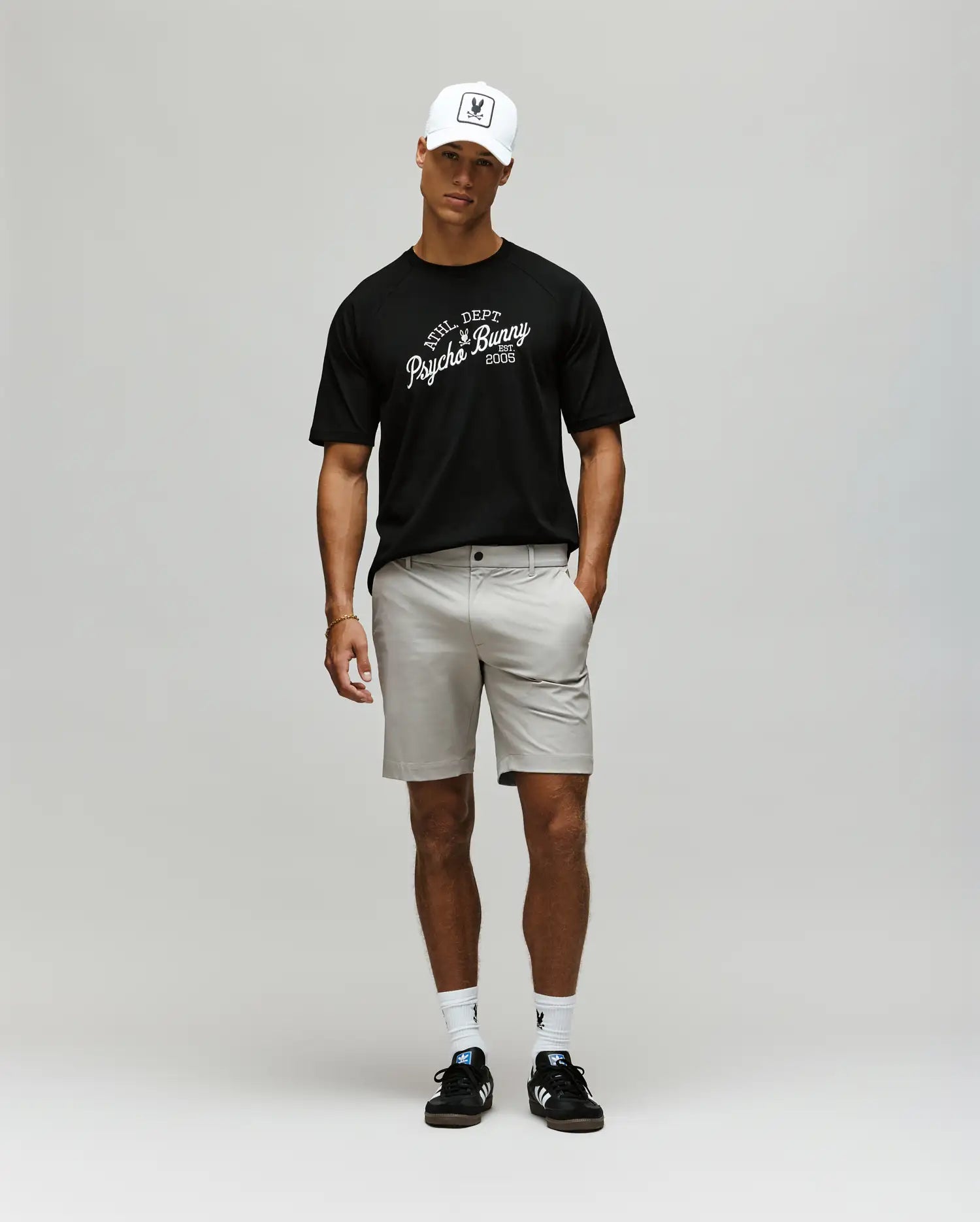 A man stands against a plain background wearing the Psycho Bunny MENS MATTEO RAGLAN GRAPHIC TEE (MT0200008), a white cap, light beige shorts, white socks, and black sandals. His arms are relaxed by his sides as he looks slightly downward.