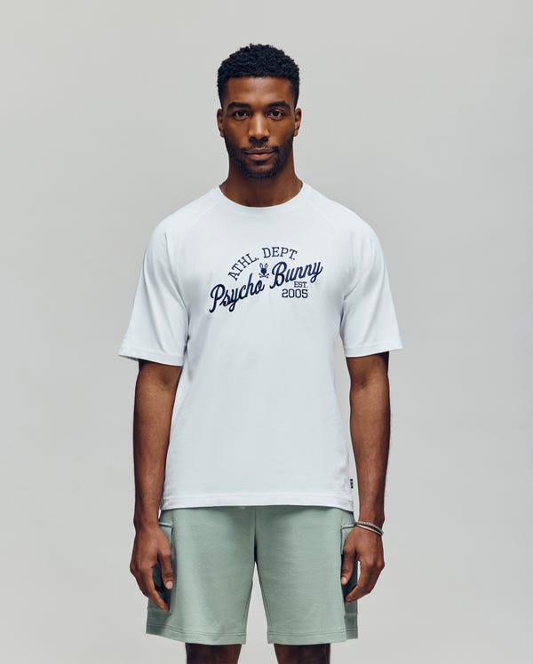 A man faces forward in a white Psycho Bunny MENS MATTEO RAGLAN GRAPHIC TEE (MT0200008) with "Psycho Bunny 2005" in navy script, paired with light green shorts, against a plain, light background—channeling classic American sporting style.
