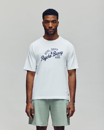 A man faces forward in a white Psycho Bunny MENS MATTEO RAGLAN GRAPHIC TEE (MT0200008) with "Psycho Bunny 2005" in navy script, paired with light green shorts, against a plain, light background—channeling classic American sporting style.