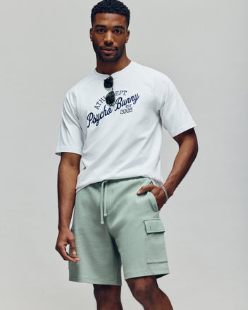 A man with a neutral expression stands against a plain background wearing the Psycho Bunny MENS MATTEO RAGLAN GRAPHIC TEE (MT0200008), light green drawstring cargo shorts, and sunglasses hanging from his collar, left hand in pocket.