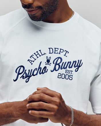 A man wears the Psycho Bunny MENS MATTEO RAGLAN GRAPHIC TEE (MT0200008), a white Pima cotton sweatshirt with blue "ATHL. DEPT. Psycho Bunny EST. 2005." text, silver bracelet, and an American sporting vibe.
