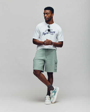 A man stands against a plain background wearing the Psycho Bunny MENS MATTEO RAGLAN GRAPHIC TEE (MT0200008), light green cargo shorts, gray socks, white sneakers, and sunglasses hanging from his shirt, capturing an American sporty vibe.
