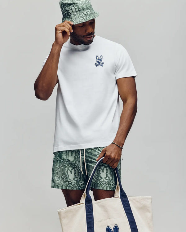 A man wears the Psycho Bunny MENS DEREK CHEST GRAPHIC TEE (MT0200009), a white Pima cotton crewneck with a small bunny print logo, along with green patterned shorts, a matching bucket hat, and holds a beige tote bag with blue handles.