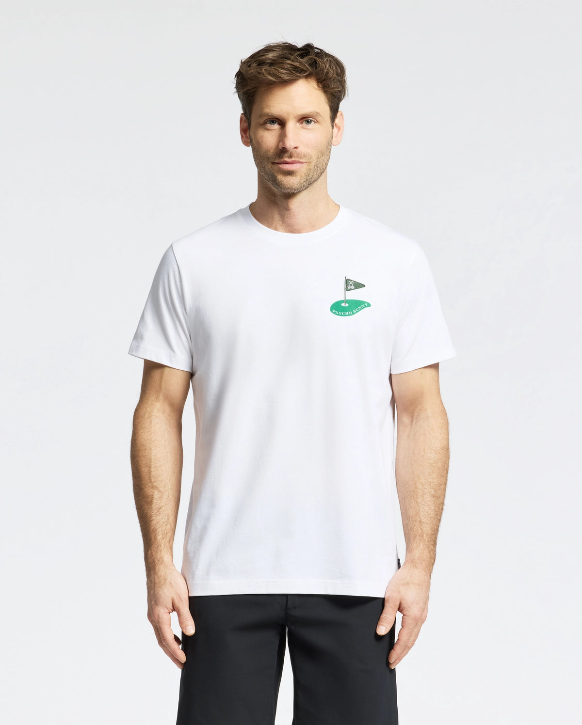 A short-haired man wears the Psycho Bunny MENS DALE GRAPHIC TEE - MT0200016, a plain white Pima cotton T-shirt with a small embroidered green logo on the chest, paired with black pants. He stands facing forward against a plain white background.