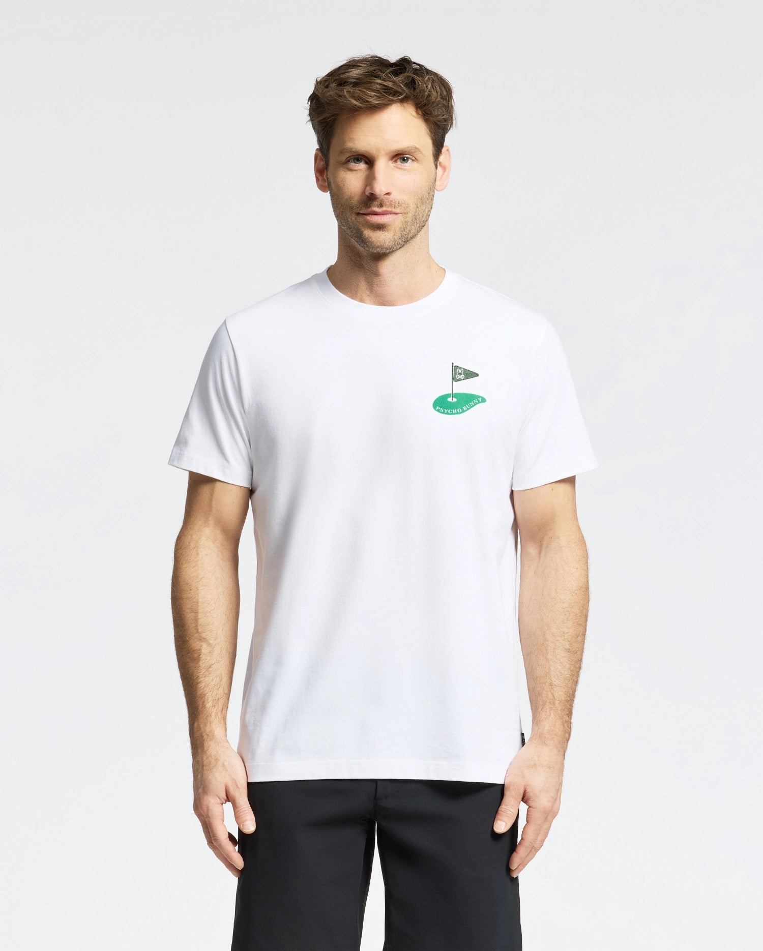 A short-haired man wears the Psycho Bunny MENS DALE GRAPHIC TEE - MT0200016, a plain white Pima cotton T-shirt with a small embroidered green logo on the chest, paired with black pants. He stands facing forward against a plain white background.