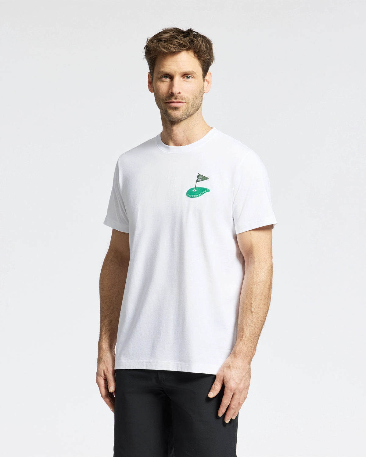 A man with short brown hair and facial hair wears a Psycho Bunny MENS DALE GRAPHIC TEE - MT0200016, a white Pima cotton T-shirt with an embroidered flag and mound logo on the chest. He stands against a plain white background, looking at the camera neutrally.