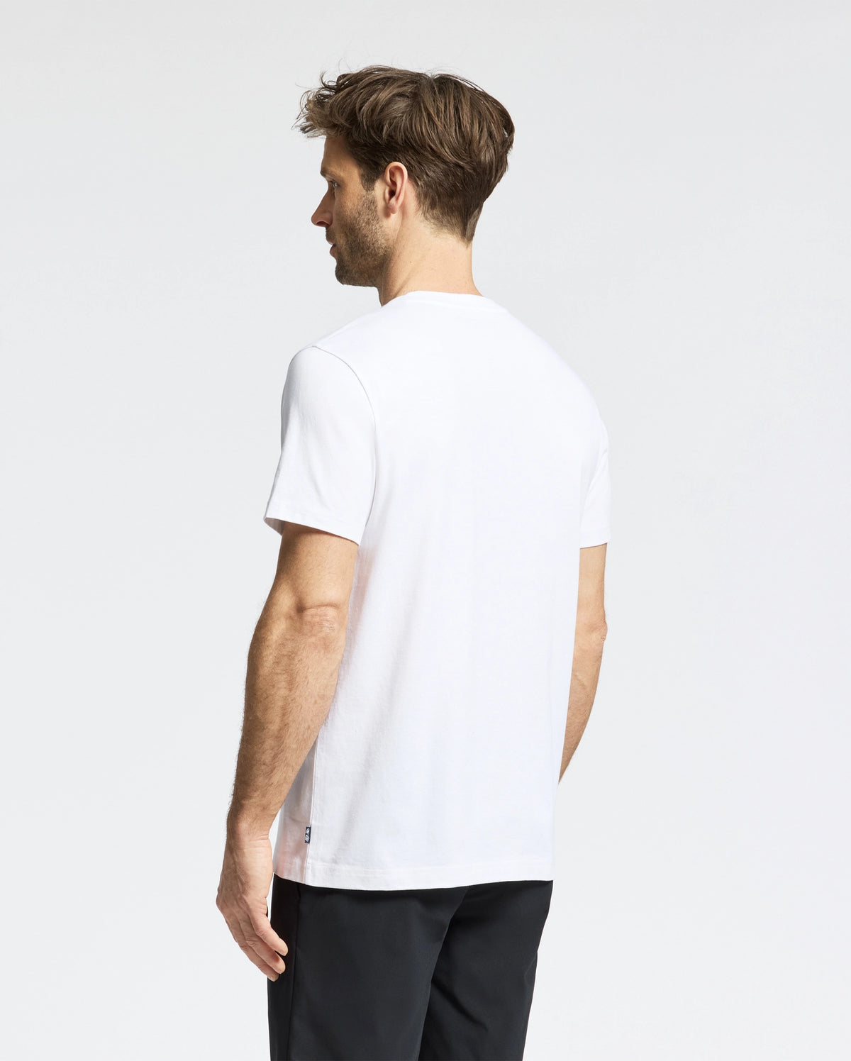 A man with short brown hair faces away, wearing a Psycho Bunny MENS DALE GRAPHIC TEE - MT0200016 and black pants. The light neutral background highlights the simplicity of his casual style.