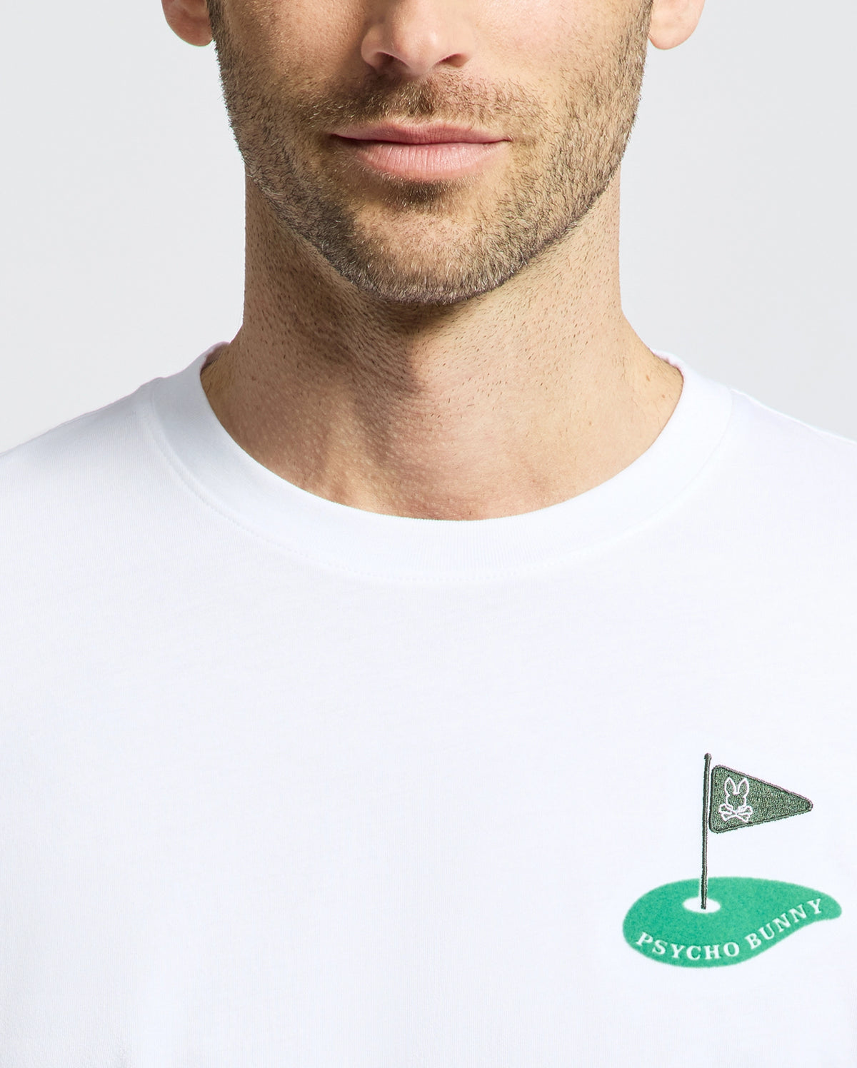 A man sports the mens Dale Graphic Tee from Psycho Bunny, featuring a small embroidered golf flag logo with "PSYCHO BUNNY" and a bunny head on the chest. His face is partially visible, showing only below the eyes.