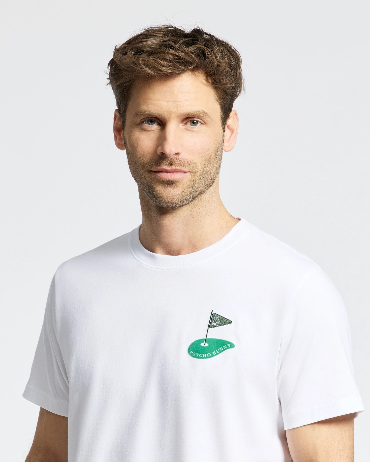 A man with short brown hair and a beard is wearing the Mens Dale Graphic Tee (MT0200016) from Psycho Bunny. It's a Pima cotton white t-shirt featuring an embroidered golf hole and flag logo on the chest, with "Psycho Bunny" text. He stands against a light gray background.