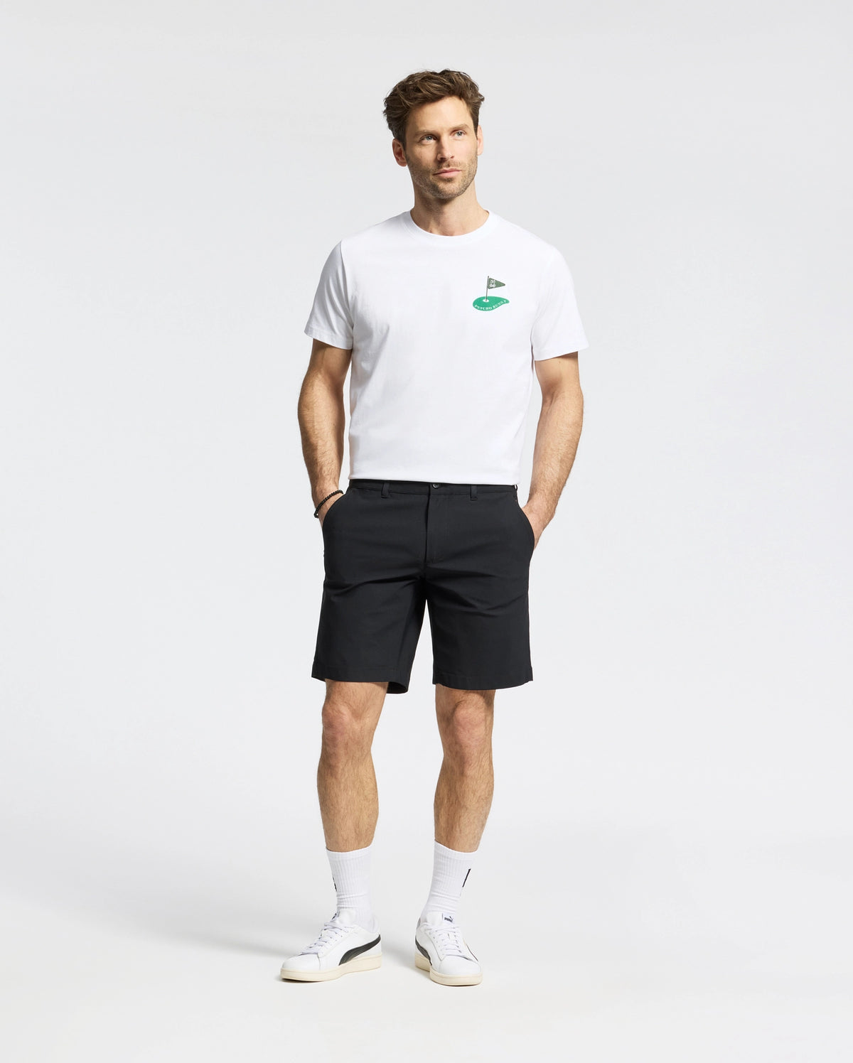 A man in the Psycho Bunny MENS DALE GRAPHIC TEE (MT0200016), a white Pima cotton T-shirt with a green embroidered logo, pairs it with black shorts. He wears white sneakers and socks, posing against a plain background with understated yet refined casual elegance.