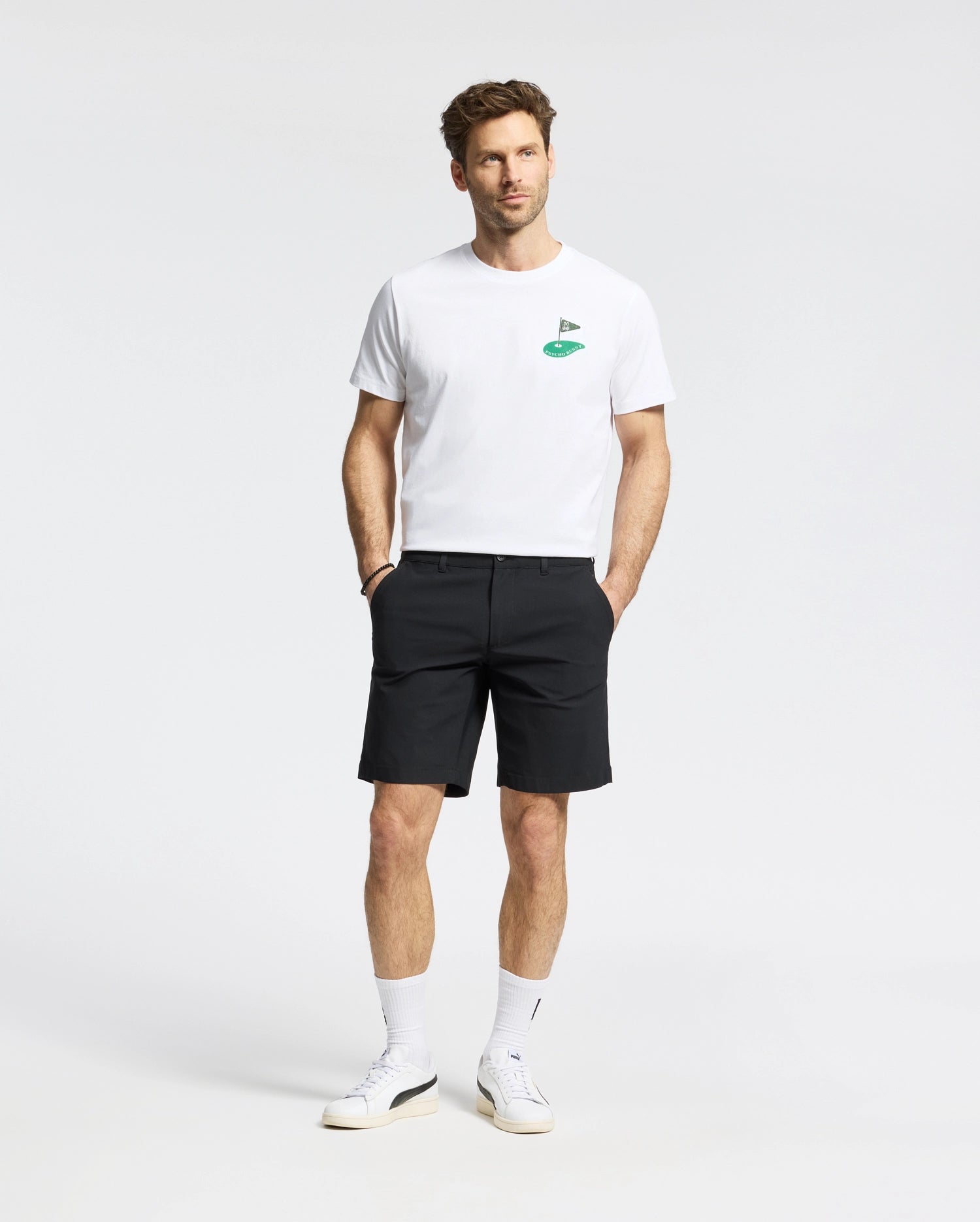 A man in the Psycho Bunny MENS DALE GRAPHIC TEE (MT0200016), a white Pima cotton T-shirt with a green embroidered logo, pairs it with black shorts. He wears white sneakers and socks, posing against a plain background with understated yet refined casual elegance.