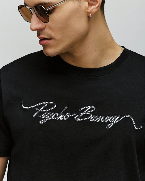 A man in profile wears the Psycho Bunny MENS CYRUS WORDMARK GRAPHIC TEE (MT0200018), a black tee featuring "Psycho Bunny" in white, cursive, rope-style lettering across the chest, and sunglasses.