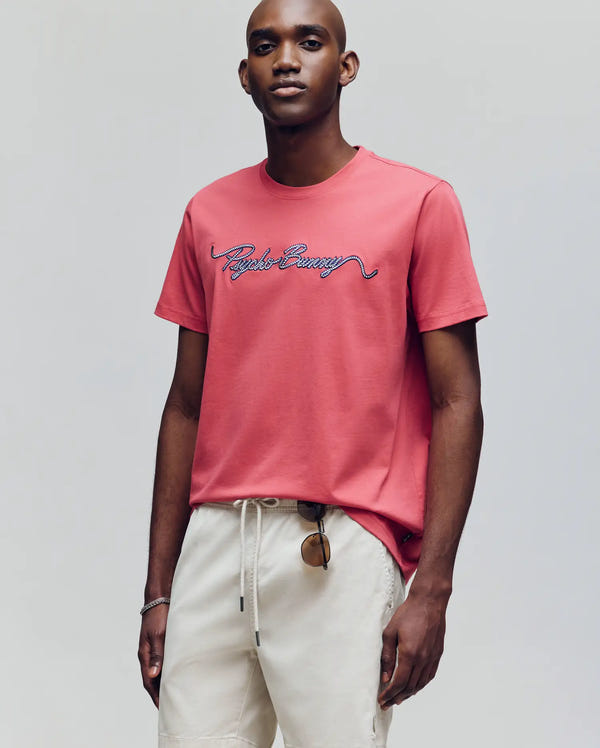 A man stands against a plain gray background, holding sunglasses by his side. He wears the Psycho Bunny MENS CYRUS WORDMARK GRAPHIC TEE (MT0200018) in pink and light beige drawstring shorts.