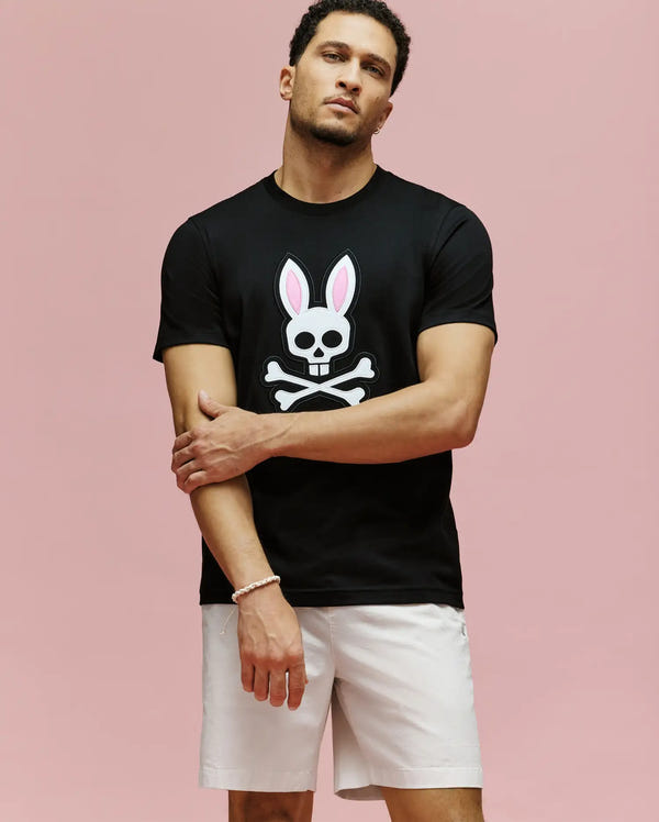 A man stands against a pink background wearing Psycho Bunny’s MENS ARCHIVE BUNNY GRAPHIC TEE—black, 100% Pima cotton from Peru, featuring an embroidered bunny skull and crossbones—paired with white shorts, his left hand on his right arm.
