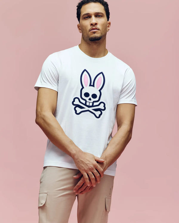 A man stands against a pink background wearing the Psycho Bunny MENS ARCHIVE BUNNY GRAPHIC TEE (MT0200031), a white Pima cotton knit crewneck with a large bunny skull and crossbones graphic, paired with light beige cargo pants.