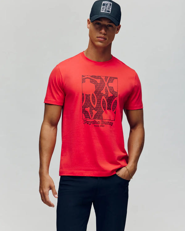 A man in a black cap, dark pants, and the Psycho Bunny MENS GLENN GRAPHIC TEE (MT0200034), featuring a bold Bunny graphic on 100% Pima cotton, stands against a plain gray background with a neutral expression.