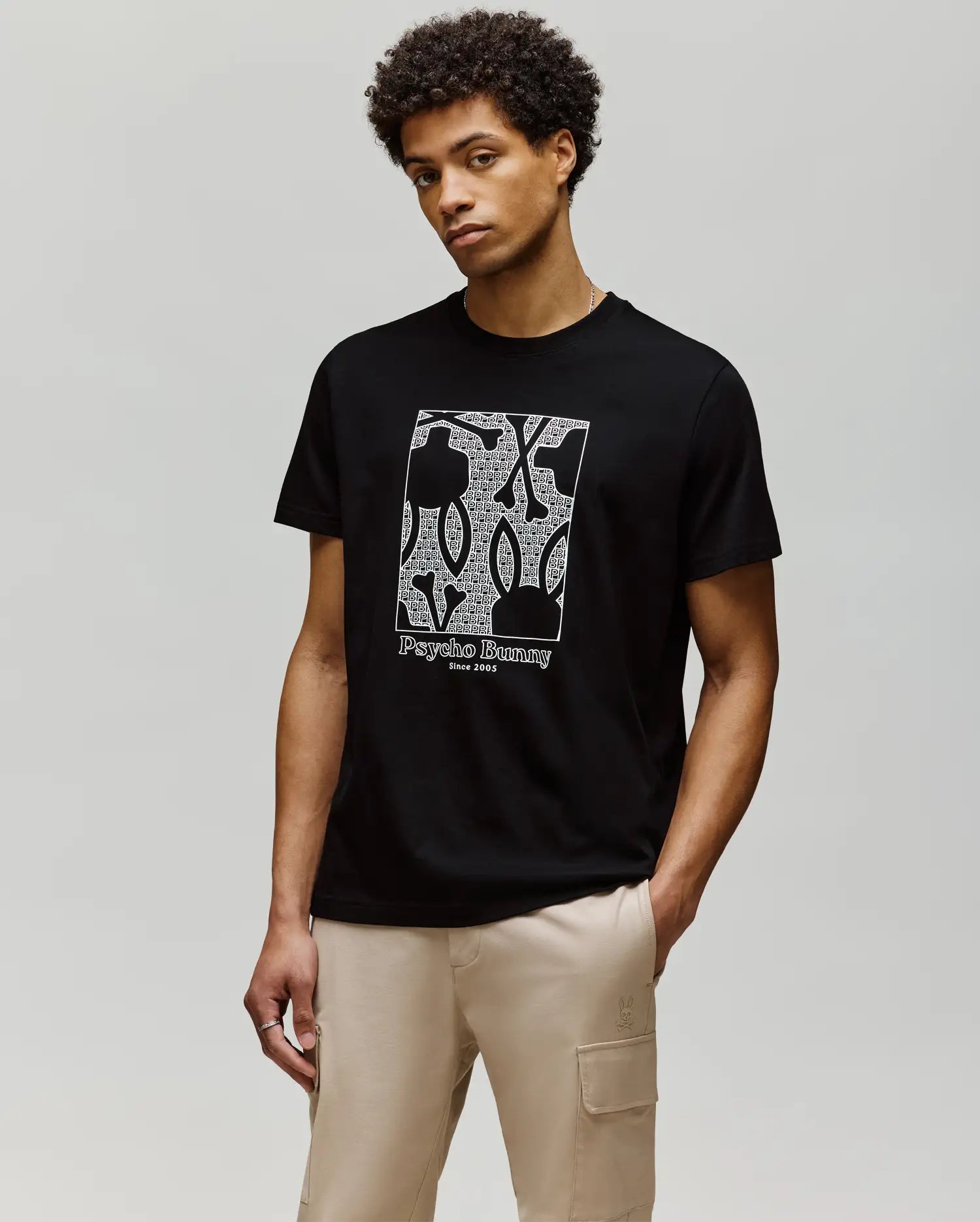 A man with short curly hair wears the Psycho Bunny MENS GLENN GRAPHIC TEE - MT0200034, a black Pima cotton T-shirt with a white Bunny graphic and "Psycho Bunny Since 2005" text, paired with beige cargo pants against a light background.