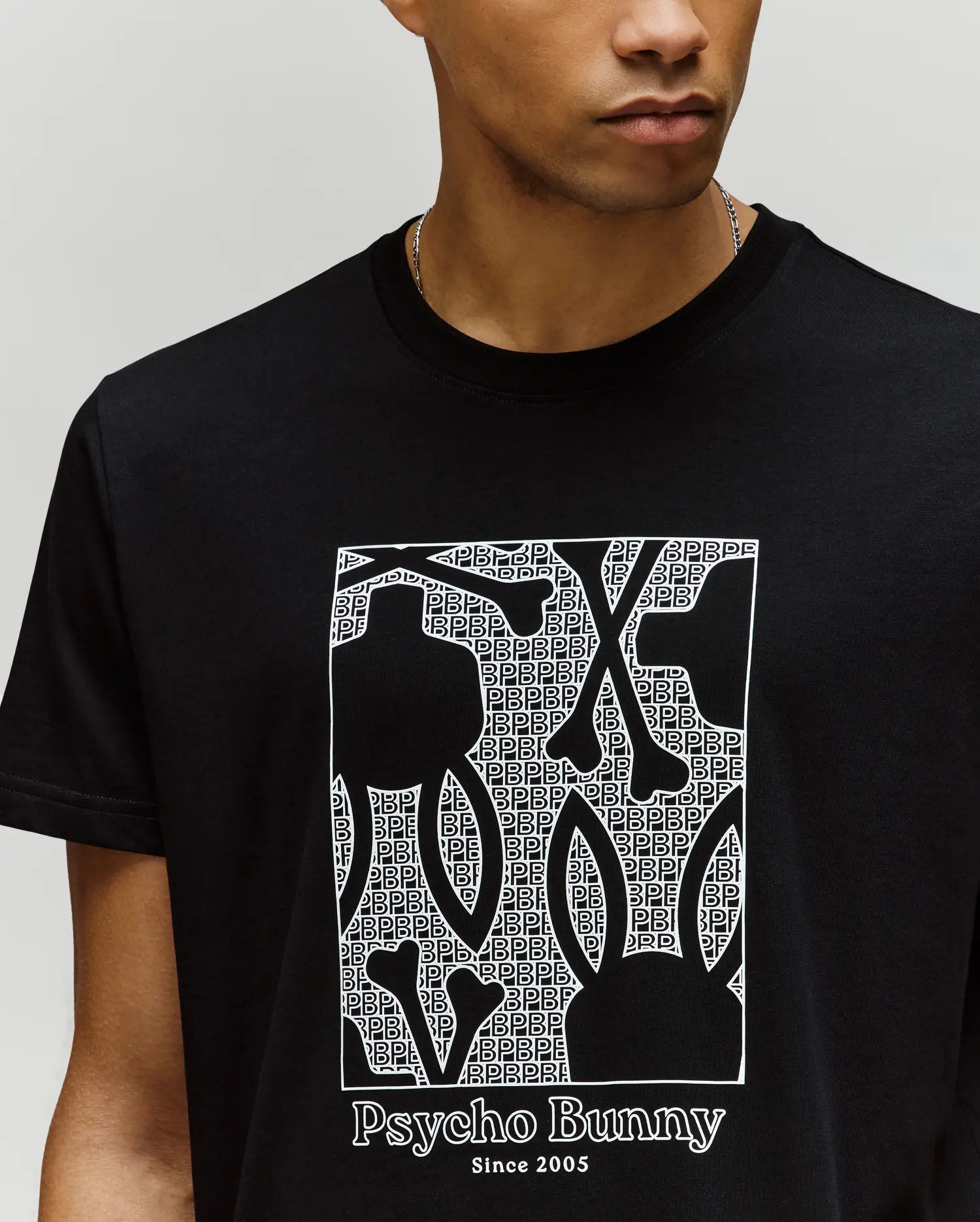 A person in a black Psycho Bunny MENS GLENN GRAPHIC TEE (MT0200034) with a bold bunny graphic filled with a repeating "PB" pattern and "Psycho Bunny Since 2005" text below.
