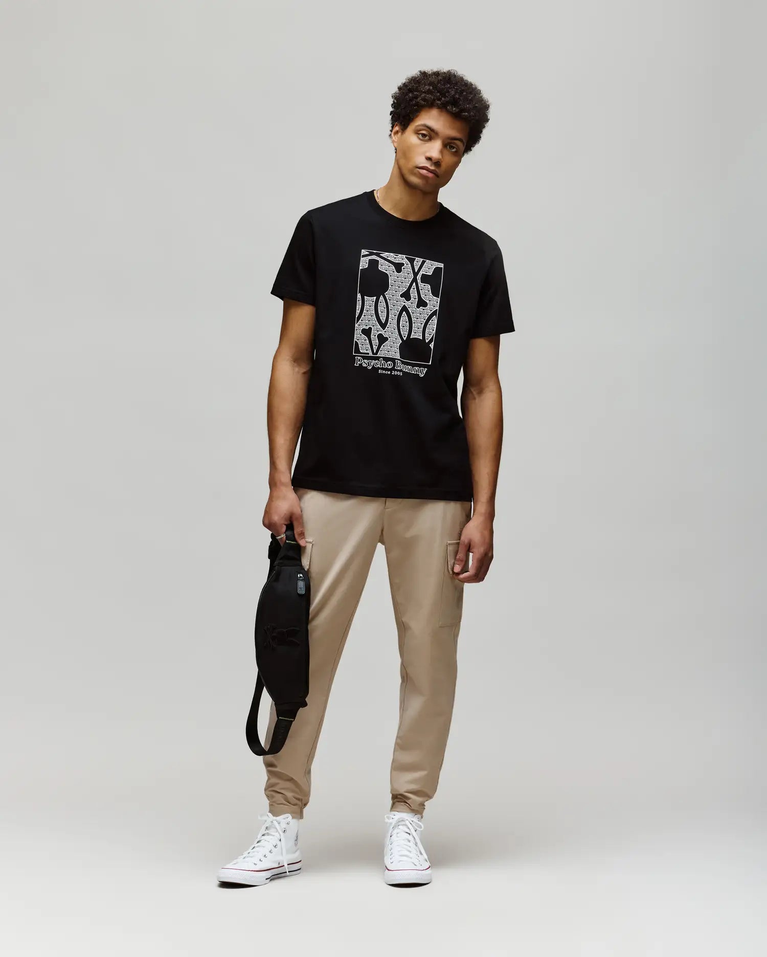 A young man stands against a plain background, wearing the Psycho Bunny MENS GLENN GRAPHIC TEE - MT0200034 in black with a Bunny graphic, paired with beige jogger pants, white high-top sneakers, and holding a black bag in his left hand.