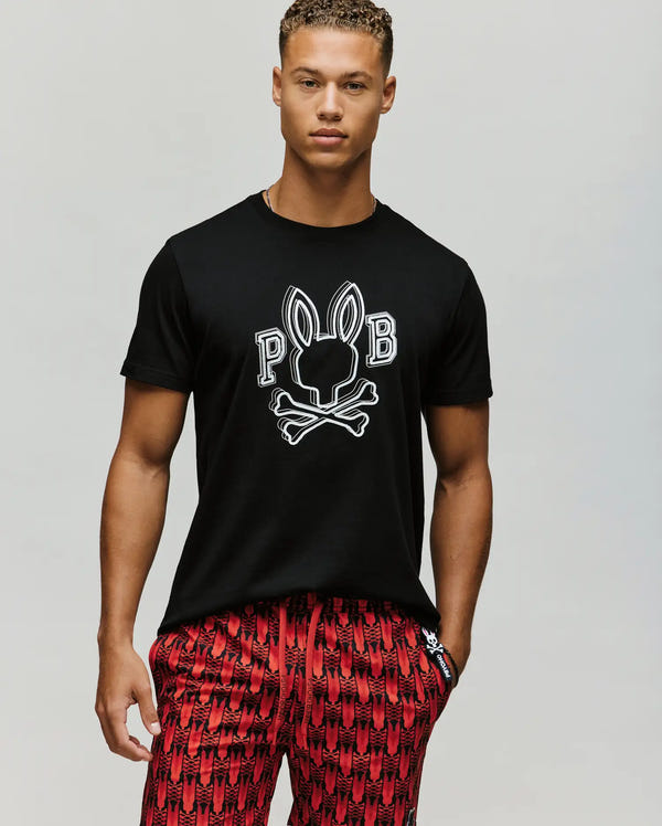 A young man wears the Psycho Bunny MENS BALES GRAPHIC TEE (MT0200035) in black, made from soft Pima cotton with a Big Bunny skull and crossbones and "P B" print, paired with red patterned shorts against a plain light background.