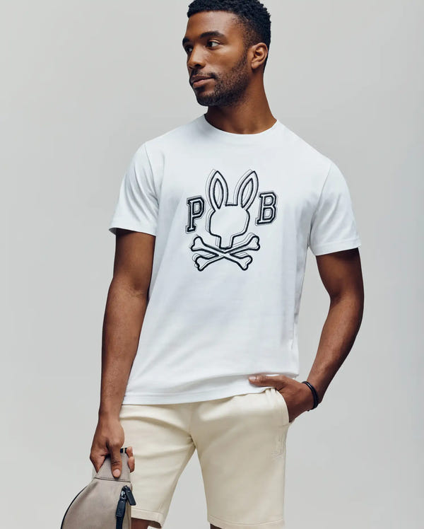 A man in a Psycho Bunny MENS BALES GRAPHIC TEE (MT0200035) with a Big Bunny print, paired with light shorts and holding a gray bag, stands against a plain light background.