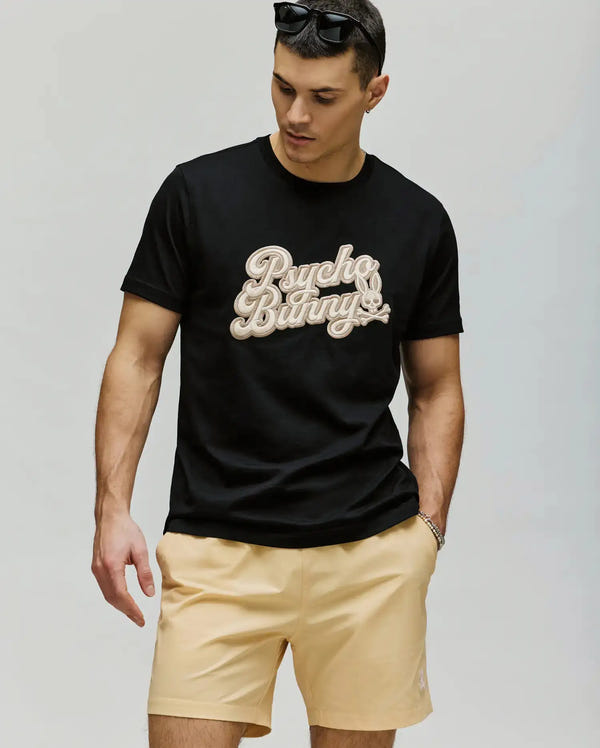 A man in a black MENS ELIAS GRAPHIC TEE by Psycho Bunny with a bold Bunny graphic and beige shorts looks down, one hand relaxed at his side, against a plain light-colored background.