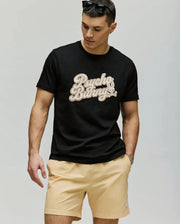 A man in a black MENS ELIAS GRAPHIC TEE by Psycho Bunny with a bold Bunny graphic and beige shorts looks down, one hand relaxed at his side, against a plain light-colored background.