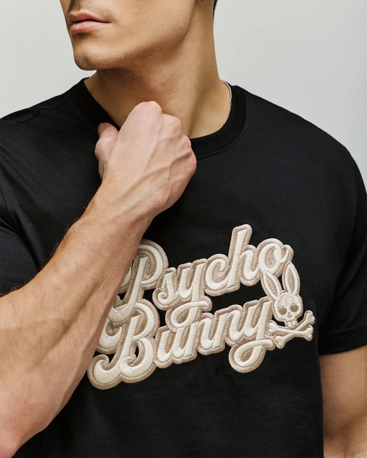 Wearing the Psycho Bunny MENS ELIAS GRAPHIC TEE, a black Pima cotton shirt with bold, textured "Psycho Bunny" lettering and a standout bunny skull logo, he showcases retro style as his hand rests near the collar.