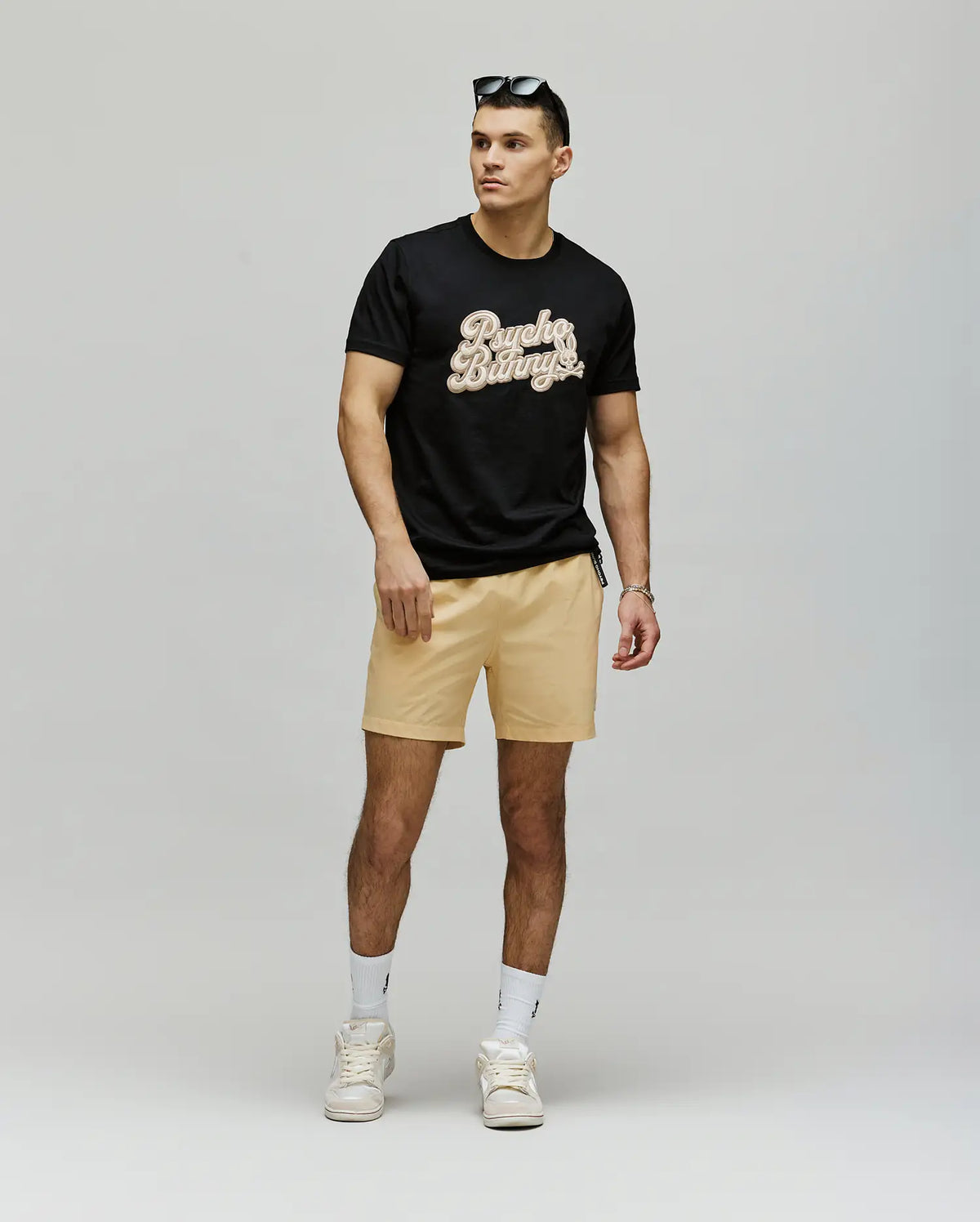 A man wears the Psycho Bunny MENS ELIAS GRAPHIC TEE (MT0200047) in black Pima cotton, paired with light beige shorts, white socks and sneakers. Sunglasses on his head, he stands against a plain background gazing to the side.