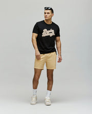 A man wears the Psycho Bunny MENS ELIAS GRAPHIC TEE (MT0200047) in black Pima cotton, paired with light beige shorts, white socks and sneakers. Sunglasses on his head, he stands against a plain background gazing to the side.