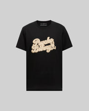 The MENS ELIAS GRAPHIC TEE (MT0200047) by Psycho Bunny features black Pima cotton, a large cream retro "Psycho Bunny" script across the chest, and a small Bunny graphic, displayed on a light gray background.