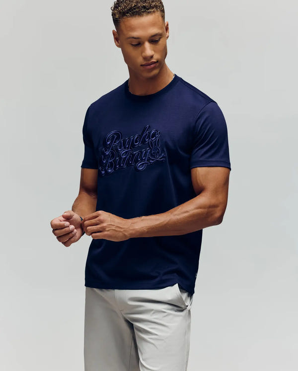 A man wearing the Psycho Bunny MENS ELIAS GRAPHIC TEE (MT0200047) in navy blue with script and Bunny graphic, paired with light gray pants, stands against a plain background looking down with his hands relaxed.