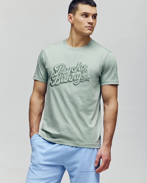 A man wearing a light green Psycho Bunny MENS ELIAS GRAPHIC TEE (MT0200047) and pale blue shorts stands with one hand in his pocket, gazing off to the side against a plain light gray background.