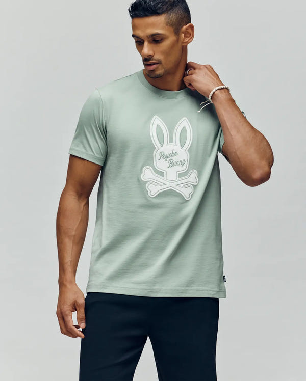 A man in the Psycho Bunny MENS DEREK CENTER GRAPHIC TEE - MT0200048, featuring a large white bunny and crossbones graphic on light green Pima cotton, pairs it with black pants against a plain light background.