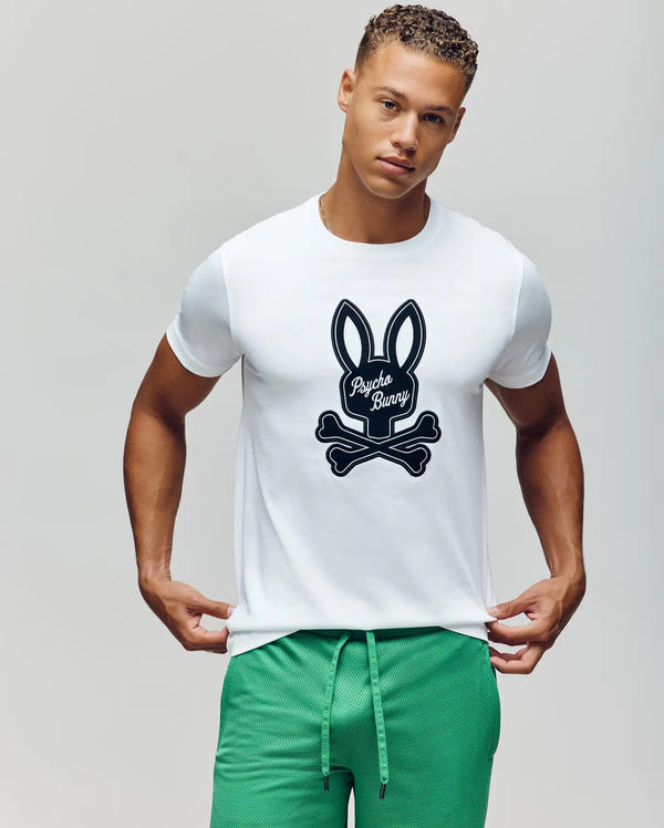 A young man with short curly hair wears the Psycho Bunny MENS DEREK CENTER GRAPHIC TEE (MT0200048), a white Pima cotton shirt with a large black bunny and crossbones, paired with green athletic shorts against a plain background.