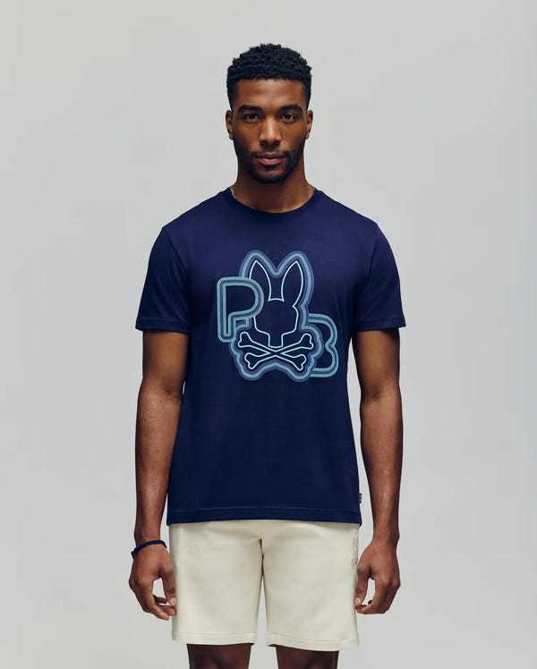 A man faces forward in the Psycho Bunny MENS CARLO GRAPHIC TEE (MT0200049), a navy blue shirt featuring a bold bunny and crossbones design, paired with light shorts, set against a plain light gray background.