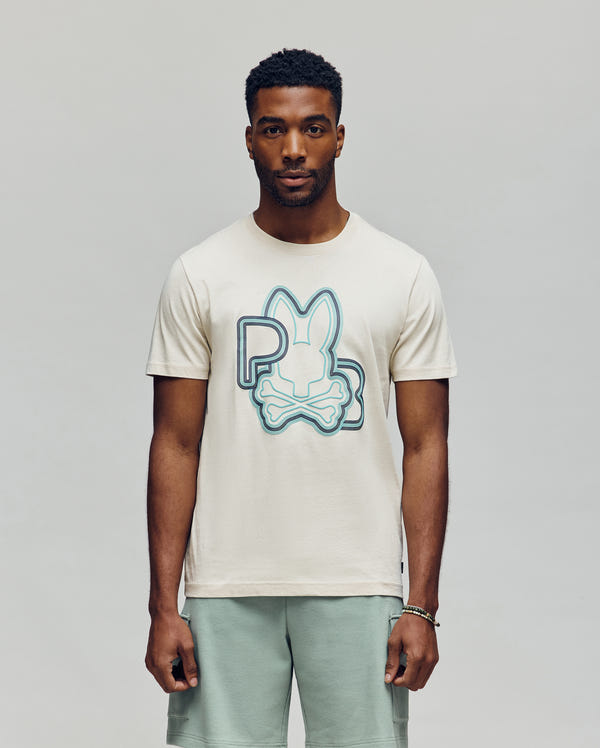 A man faces forward in Psycho Bunny’s MENS CARLO GRAPHIC TEE - MT0200049, a cream Pima cotton jersey with a large blue and aqua stylized bunny and bones, paired with light green shorts and a bracelet, set against a plain light background.