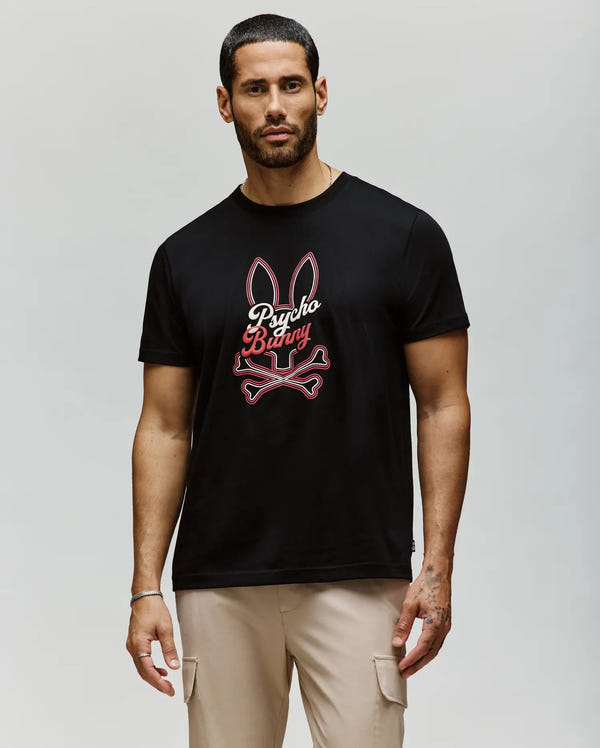 A man with short dark hair and a beard wears the Psycho Bunny MENS GARRET GRAPHIC TEE (MT0200050), a black shirt featuring bunny ears, crossbones, and "Psycho Bunny" in red and white, paired with beige pants against a light background.
