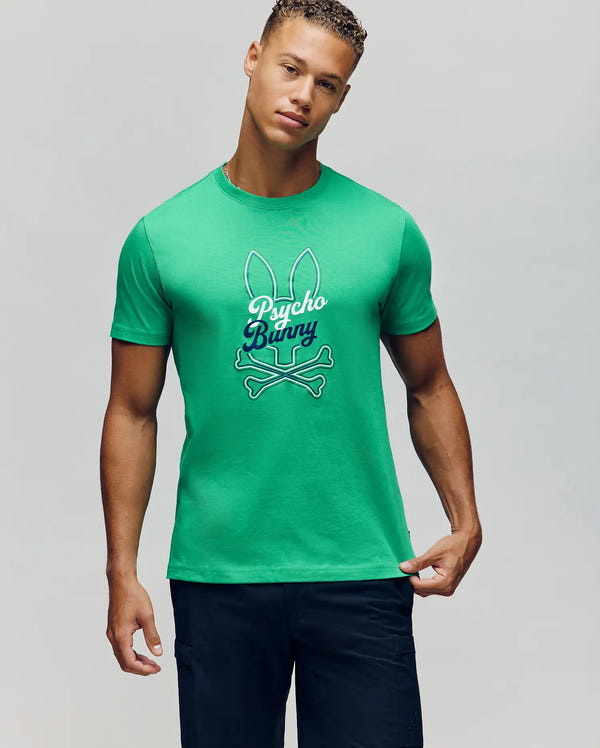 A man wearing the Psycho Bunny MENS GARRET GRAPHIC TEE - MT0200050, a green 100% Pima cotton shirt with a Big Bunny graphic and "Psycho Bunny" text, paired with dark shorts, stands against a plain gray background.