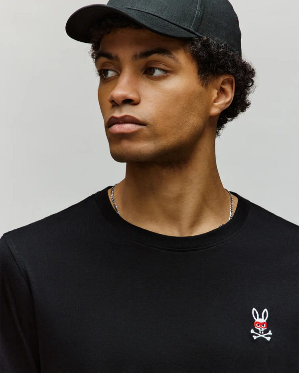 A young man in a black Psycho Bunny MENS MISCHIEF BUNNY TEE (MT0200051), crafted from soft Pima cotton and featuring an embroidered Mischief Bunny logo, looks to his left against a plain background.