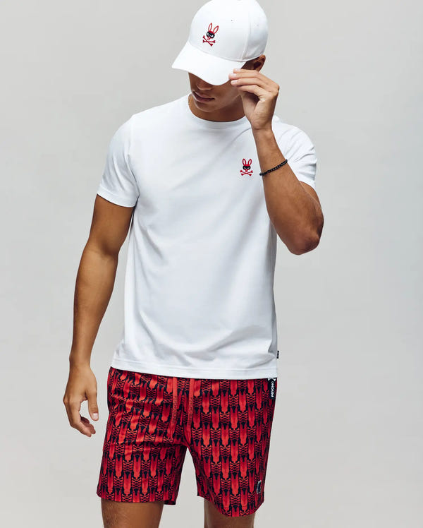 A man in a Psycho Bunny MENS MISCHIEF BUNNY TEE (MT0200051), white with a red bunny logo, pairs it with a matching cap and red patterned shorts, standing against a plain backdrop while tilting his cap to partially cover his face.