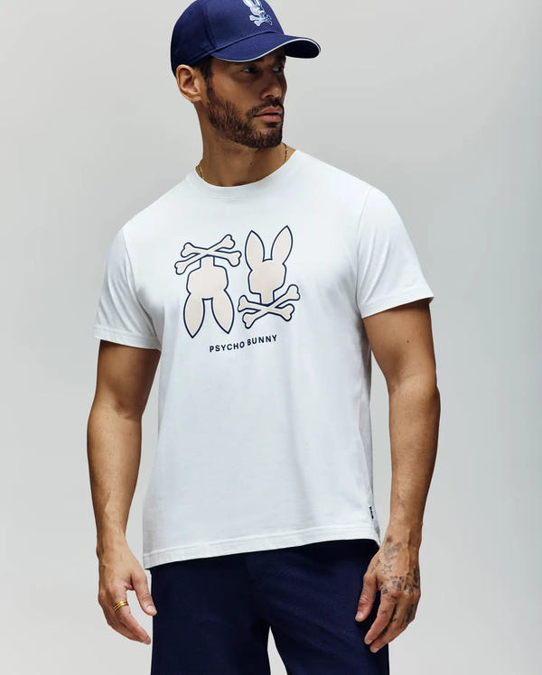 A man in a blue cap, navy pants, and a Psycho Bunny MENS DAWSON GRAPHIC TEE (MT0200053) made from soft Pima cotton with the iconic Bunny logo stands against a light background, looking to his left.