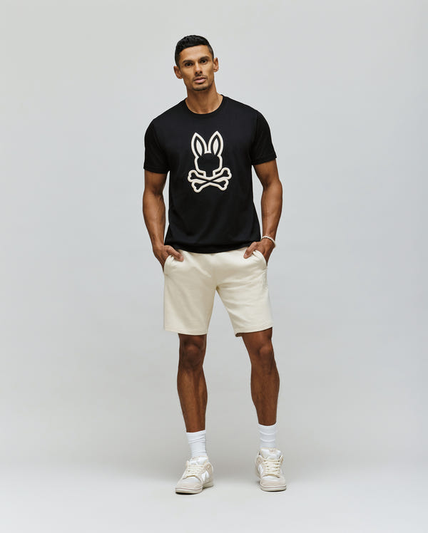 A man stands against a plain background wearing the Psycho Bunny MENS NICHOLAS EMBROIDERED TEE - MT0200054, cream shorts, white crew socks, and white sneakers, with his hands in his pockets.
