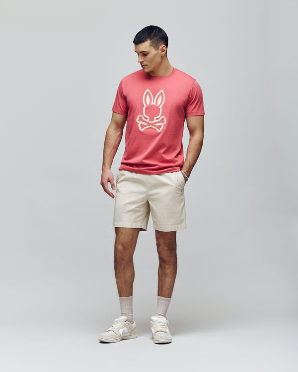 A man stands against a plain background wearing the Psycho Bunny MENS NICHOLAS EMBROIDERED TEE (MT0200054) in coral, paired with beige shorts, socks, and sneakers. He has one hand in his pocket and looks slightly to the side.