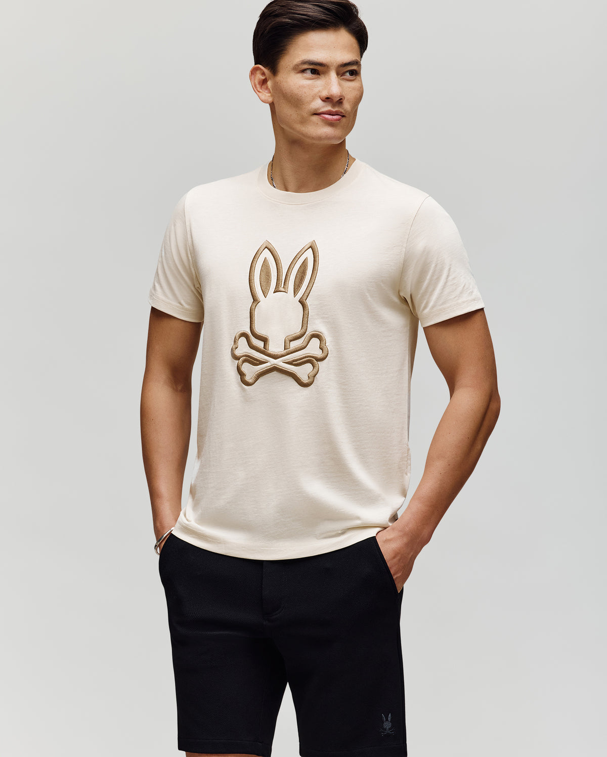 A man wears the Psycho Bunny MENS NICHOLAS EMBROIDERED TEE (MT0200054), featuring a large bunny and crossbones graphic, paired with black shorts, standing with hands in pockets against a plain background.