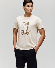 A man wears the Psycho Bunny MENS NICHOLAS EMBROIDERED TEE (MT0200054), featuring a large bunny and crossbones graphic, paired with black shorts, standing with hands in pockets against a plain background.
