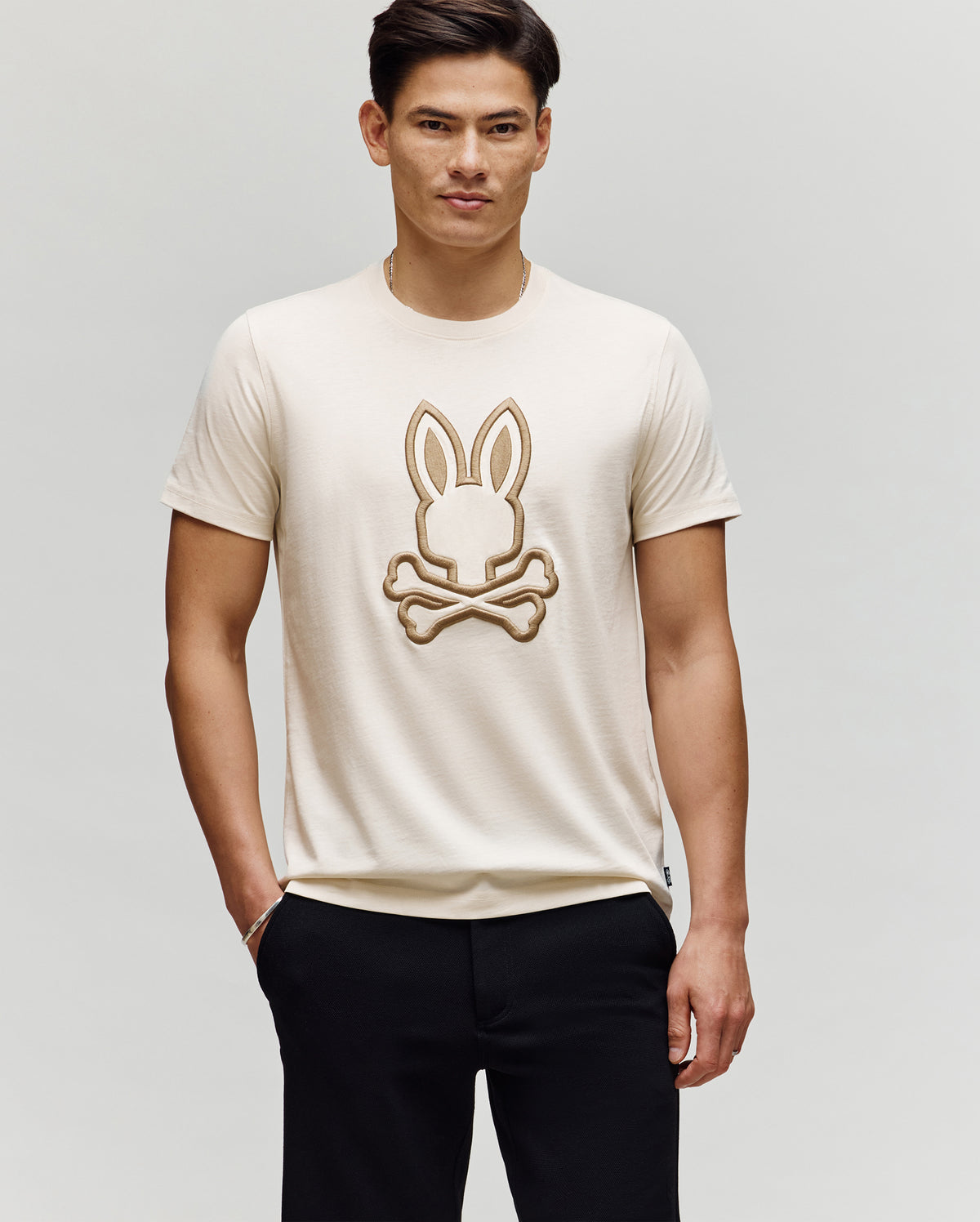 A man with short dark hair models the Psycho Bunny MENS NICHOLAS EMBROIDERED TEE (MT0200054), a cream-colored shirt with an outlined bunny head above crossed bones, standing with one hand in his pocket against a plain background.
