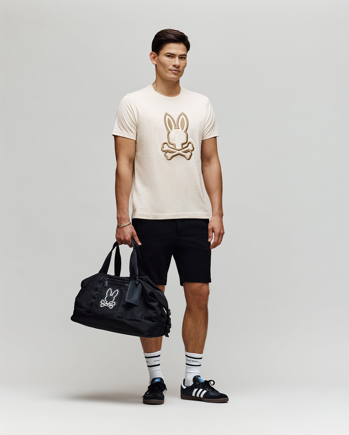 A man stands against a plain background wearing the Psycho Bunny MENS NICHOLAS EMBROIDERED TEE (MT0200054), black shorts, striped white socks, black Adidas sneakers, and holding a black duffel bag with a matching bunny logo.